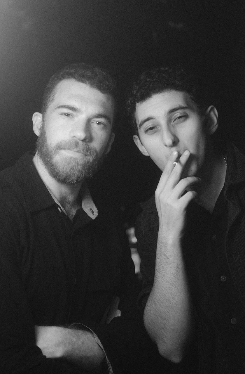 Two men at night, one drawing on a cigarette, close black-and-white double portrait
