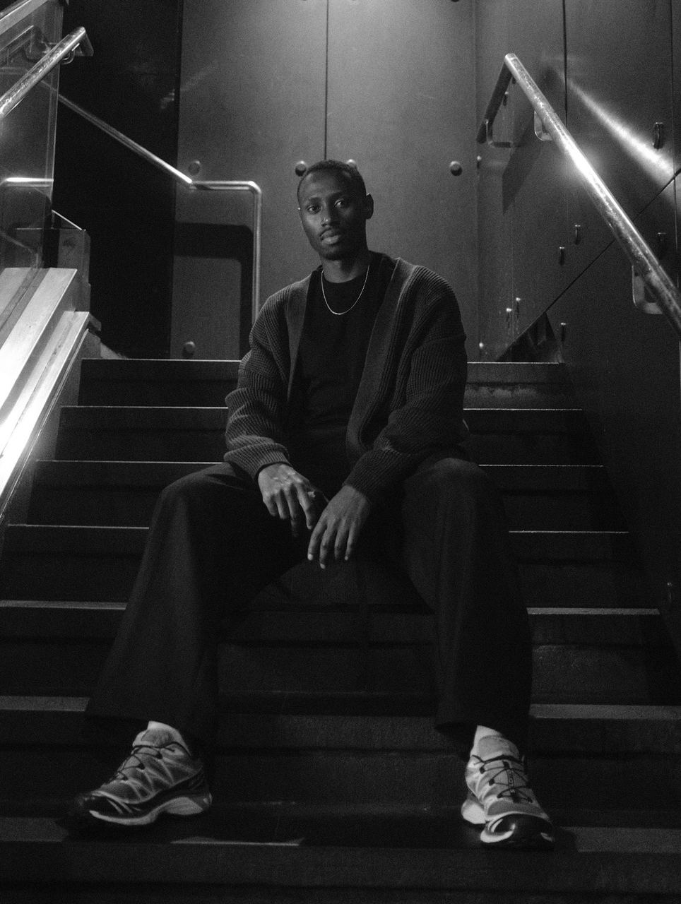 A man in a cardigan seated on a dim staircase, full-length black-and-white portrait