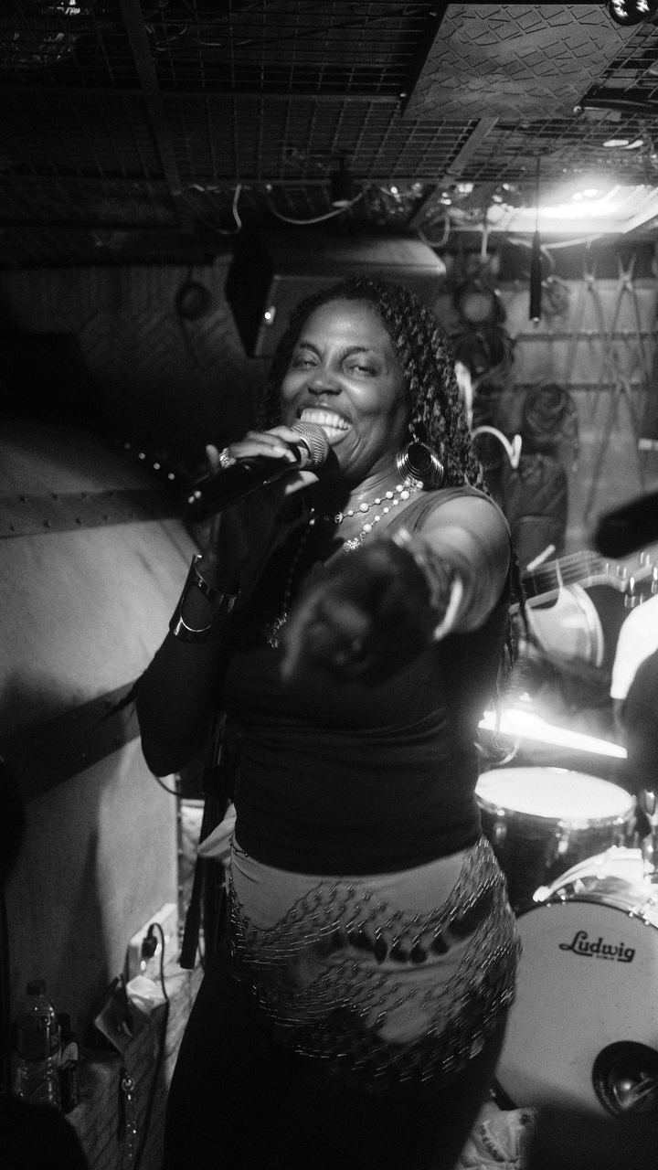 A singer laughing into the mic and pointing to the crowd at a small live show, black and white