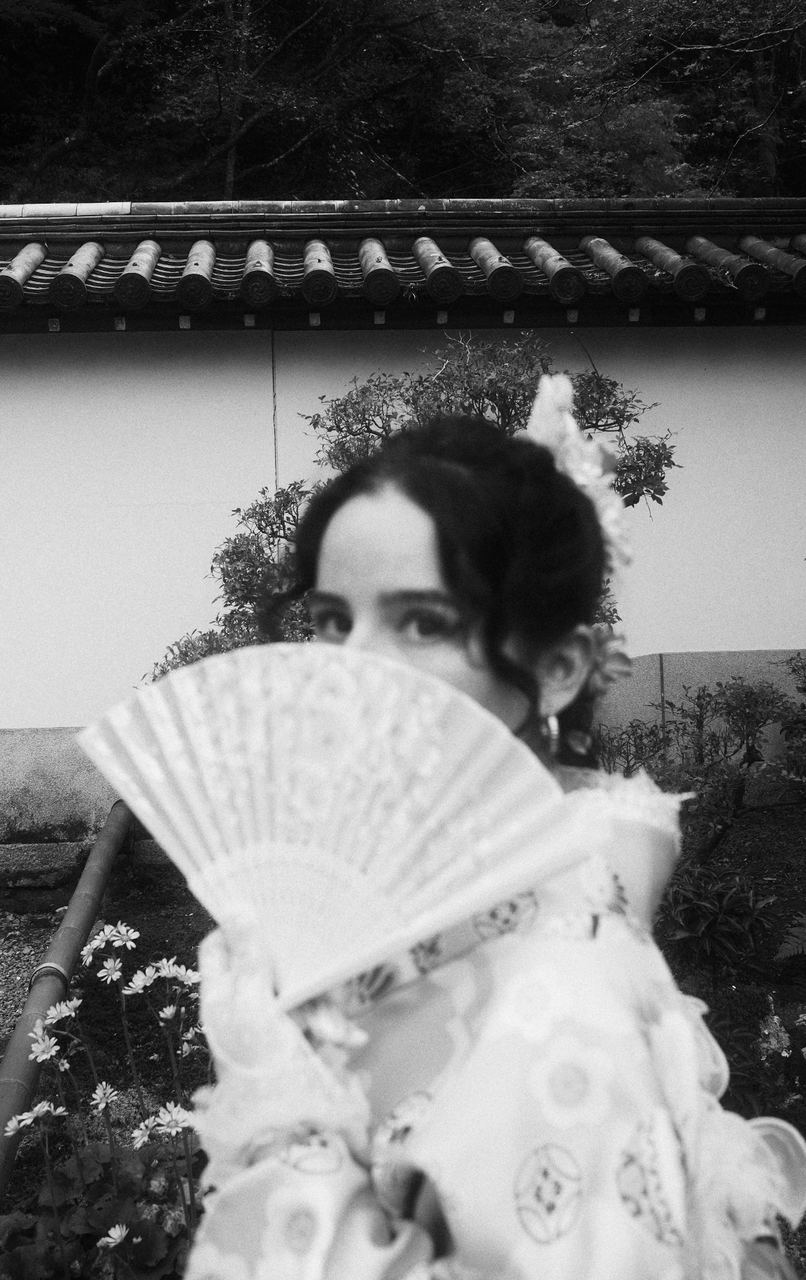 A woman in a kimono holding a lace fan before a temple wall and garden, black and white, Kyoto, Japan