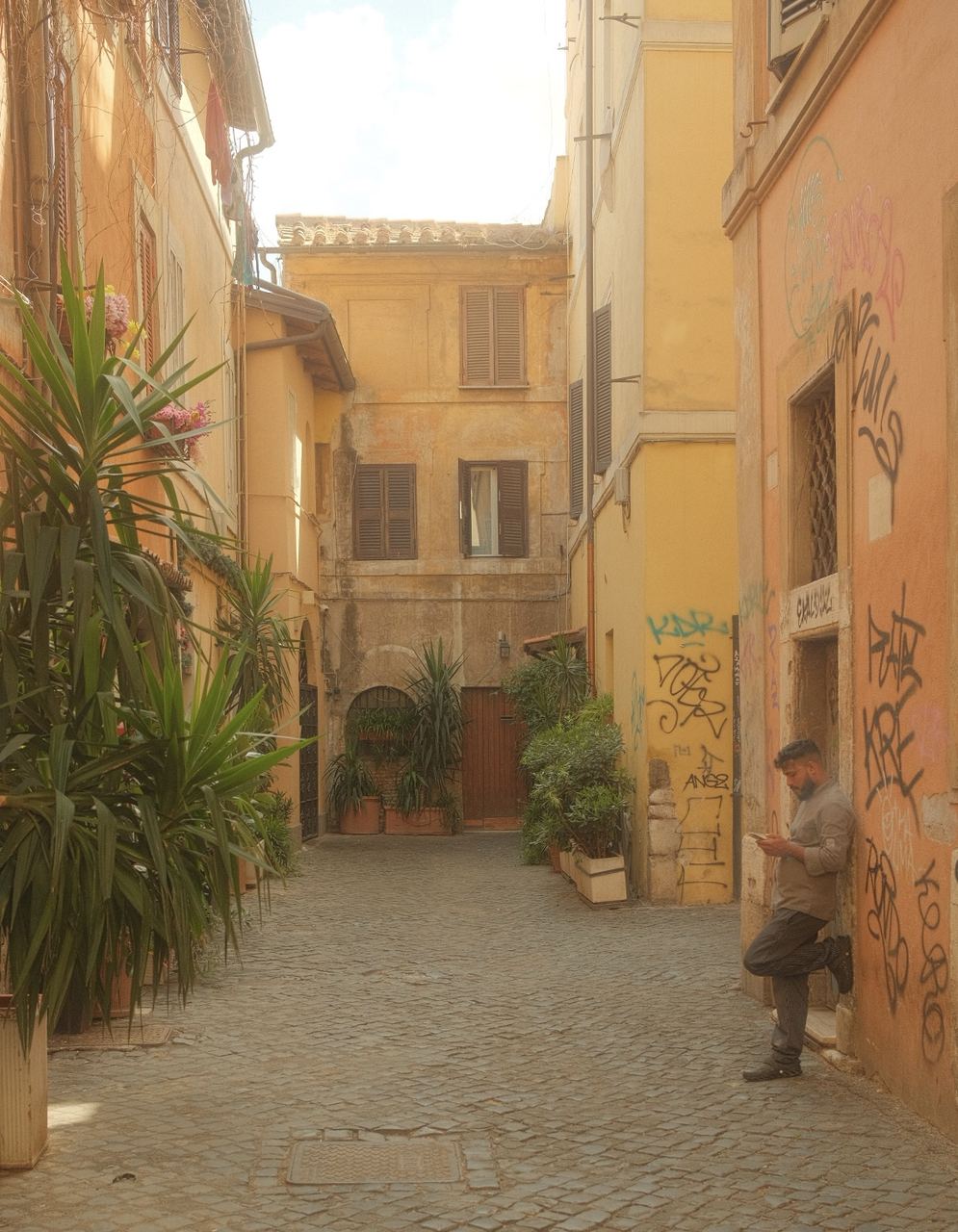 A man checks his phone against a graffiti wall in a plant-filled Trastevere alley, Rome