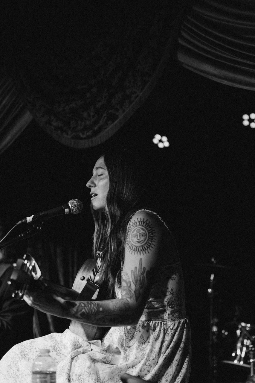 A seated musician with a tattooed arm singing into the mic, black and white