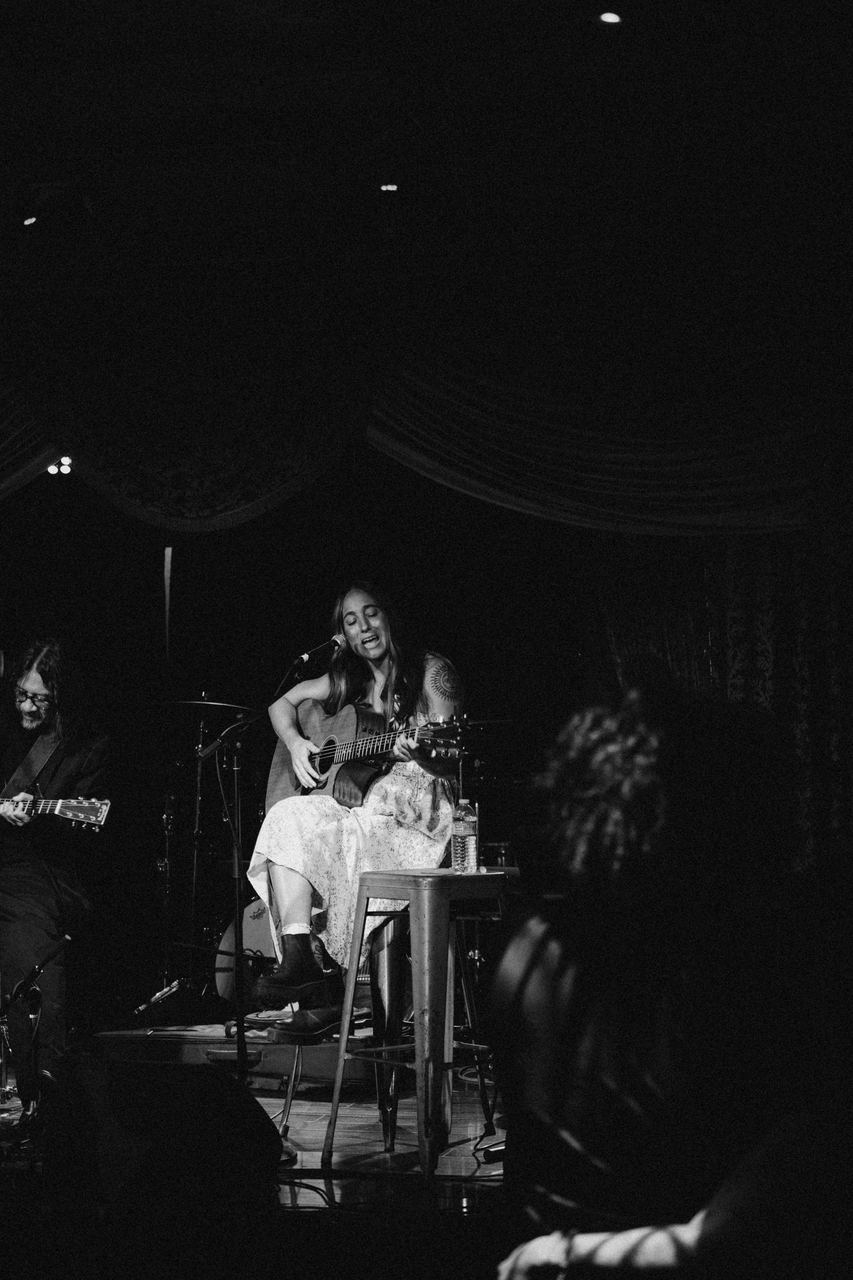 Wide view of a musician performing on a dim stage with her band, black and white